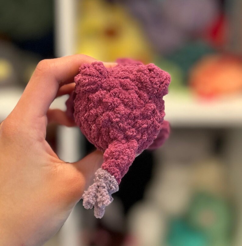May include: A plush purple and lavender crocheted toy with a textured surface. The toy is shaped like a small animal with a tail.