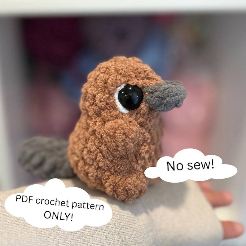 May include: A brown and gray crocheted plush toy shaped like a platypus. The toy has large black eyes and a small gray beak. The toy is sitting on a person's hand. The text "No sew!" is written in a white speech bubble above the toy. The text "PDF crochet pattern ONLY!" is written in a white speech bubble below the toy.