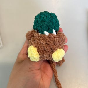 Op de afbeelding: Een handgemaakt bruin en groen pluche eendenspeelgoed met gele voetjes. De eend heeft een zachte textuur en een lange, bruine pijpenragerstaart. Het speelgoed is ontworpen om zacht en knuffelig te zijn.