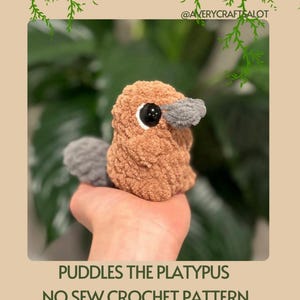 May include: A crocheted platypus toy with brown and gray fur. The toy is being held in a hand. The text "Puddles the Platypus No Sew Crochet Pattern with Low Sew Option" is at the bottom of the image.