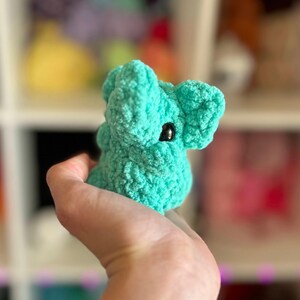 May include: A small, teal blue crocheted stuffed animal shaped like an elephant. The elephant has a single black eye and a textured surface.