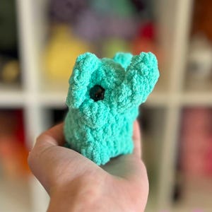 May include: A small, teal blue crocheted elephant toy with black eyes. The elephant is being held in a person's hand.