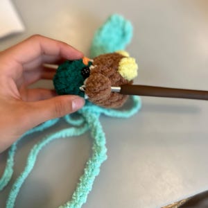 Op de afbeelding: Een close-up van een handgemaakte gehaakte eend met bruine, groene en gele details. De eend wordt gehaakt met een bruine haaknaald. De eend heeft een blauwgroene vleugel en blauwgroen garen is bevestigd.
