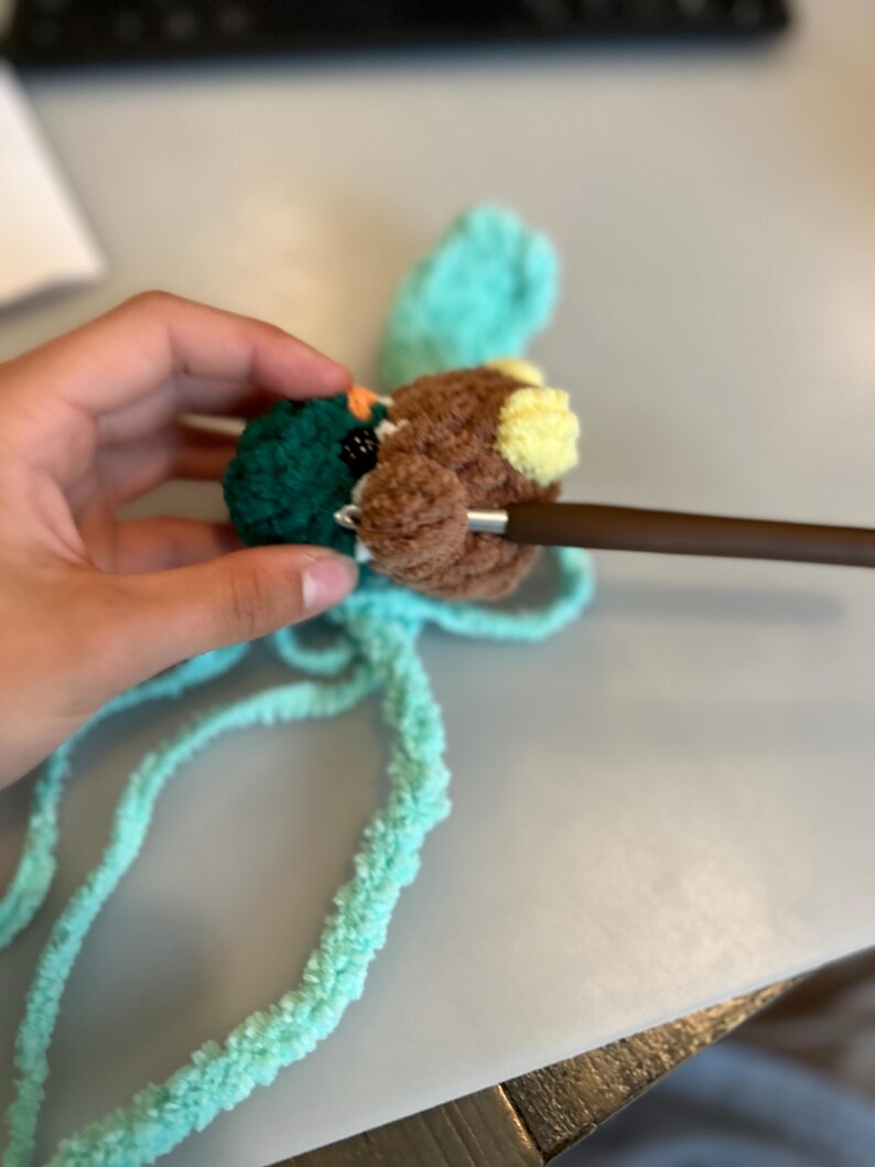 Op de afbeelding: Een close-up van een handgemaakte gehaakte eend met bruine, groene en gele details. De eend wordt gehaakt met een bruine haaknaald. De eend heeft een blauwgroene vleugel en blauwgroen garen is bevestigd.