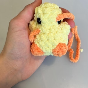 Op de afbeelding: Een handgemaakt, pluche eendenspeelgoed in geel en oranje. De eend heeft zwarte ogen, oranje voeten en een bijpassende snavel. Het speelgoed is gemaakt van zacht, fuzzy garen en wordt in een hand gehouden.