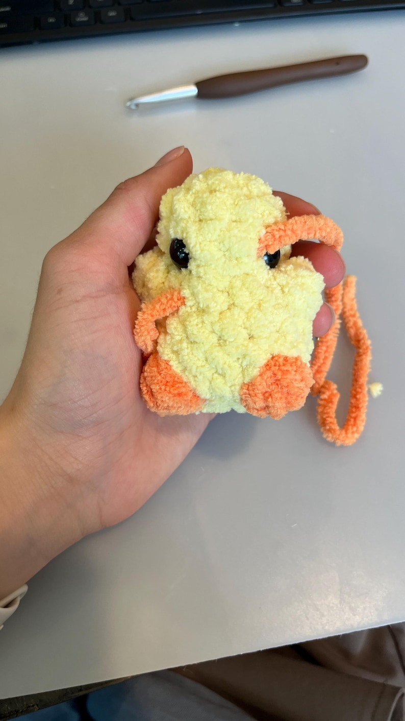 Op de afbeelding: Een handgemaakt, pluche eendenspeelgoed in geel en oranje. De eend heeft zwarte ogen, oranje voeten en een bijpassende snavel. Het speelgoed is gemaakt van zacht, fuzzy garen en wordt in een hand gehouden.