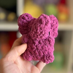 May include: A small, crocheted elephant plush toy in a deep pink color. The toy has a textured, fuzzy surface and is made with a soft yarn.