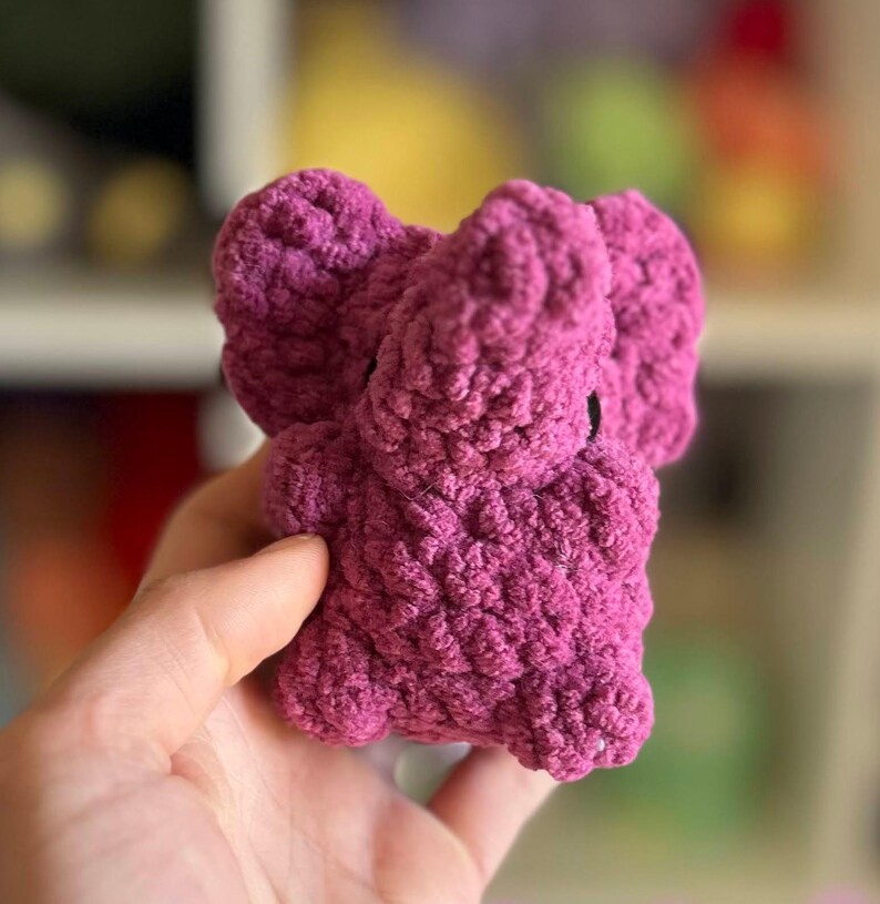 May include: A small, crocheted elephant plush toy in a deep pink color. The toy has a textured, fuzzy surface and is made with a soft yarn.