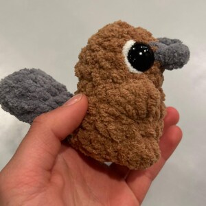 May include: A brown and gray crocheted plush toy shaped like a platypus. The toy has a small, black eye and a gray beak.