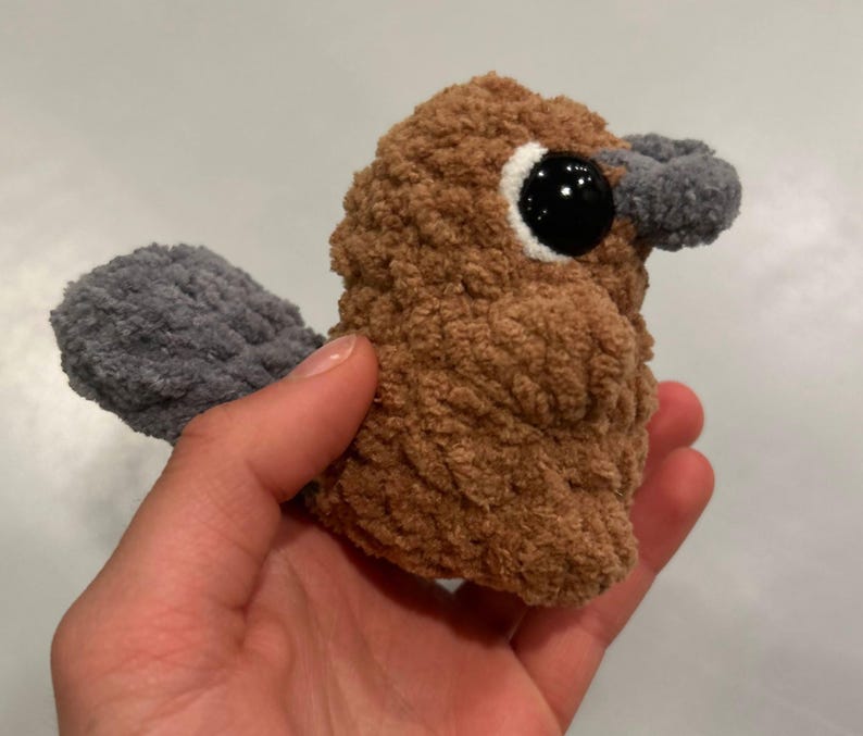 May include: A brown and gray crocheted plush toy shaped like a platypus. The toy has a small, black eye and a gray beak.