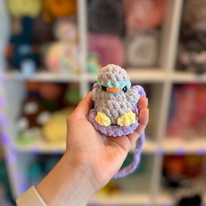 Op de afbeelding: Een handgemaakte gehaakte eend in grijstinten, blauw, geel en paars. De eend heeft een oranje snavel en wordt in een hand gehouden. De achtergrond toont planken gevuld met garen.