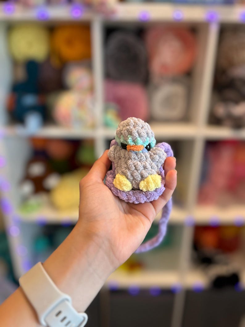 Op de afbeelding: Een handgemaakte gehaakte eend in grijstinten, blauw, geel en paars. De eend heeft een oranje snavel en wordt in een hand gehouden. De achtergrond toont planken gevuld met garen.