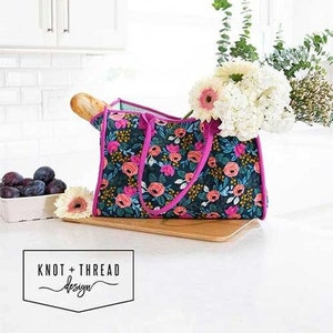 May include: A floral tote bag with pink handles and trim, filled with flowers and a baguette. The bag has a dark background with pink and orange flowers. The text "ALL THE THINGS TOTE" is above the bag.