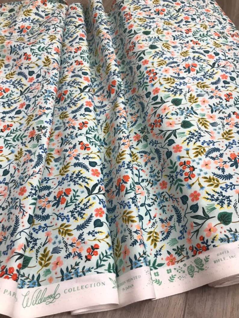Peut inclure: Un rouleau de tissu avec un motif floral de la collection Wildwood. Le motif pr&eacute;sente de petites fleurs orange, roses et bleues, ainsi que du feuillage vert et dor&eacute;, sur un fond bleu clair. Le tissu est s&eacute;rigraphi&eacute; au Japon.