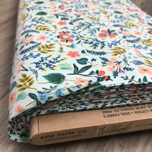 Peut inclure: Rouleau de tissu &agrave; motifs floraux. Le tissu pr&eacute;sente un fond bleu clair orn&eacute; de fleurs orange, bleues et jaunes, et de feuillage vert. L'&eacute;tiquette indique "Rifle Paper Co. For Cotton + Steel".