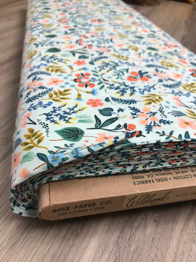 Peut inclure: Rouleau de tissu &agrave; motifs floraux. Le tissu pr&eacute;sente un fond bleu clair orn&eacute; de fleurs orange, bleues et jaunes, et de feuillage vert. L'&eacute;tiquette indique "Rifle Paper Co. For Cotton + Steel".