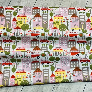 May include: White fabric with a repeating pattern of colorful houses, trees, and cars. The houses are pink, yellow, and gray with red roofs. The trees are green and the cars are red. The background is white.