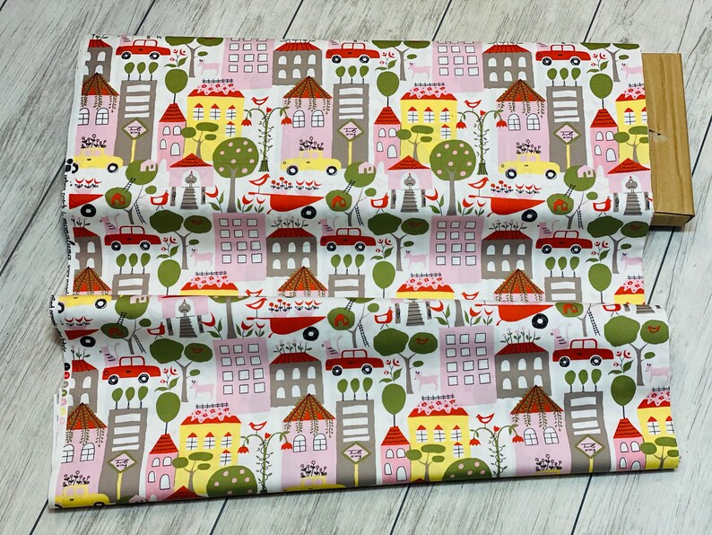 May include: White fabric with a repeating pattern of colorful houses, trees, and cars. The houses are pink, yellow, and gray with red roofs. The trees are green and the cars are red. The background is white.