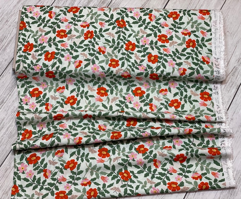 K&ouml;nnte beinhalten: Ein wei&szlig;er Stoff mit einem sich wiederholenden Muster aus roten und rosa Bl&uuml;ten, gr&uuml;nen Bl&auml;ttern und kleinen Schmetterlingen. Der Stoff hat einen wei&szlig;en Spitzenbesatz am Rand.