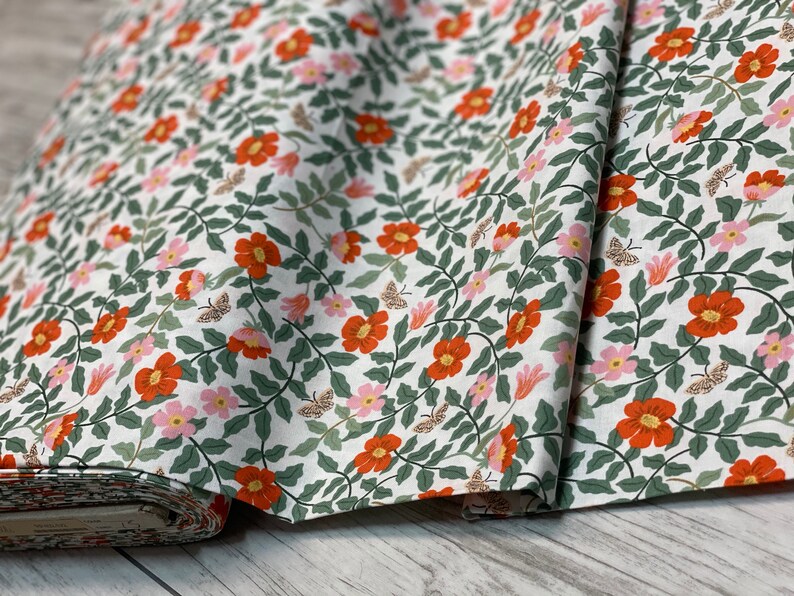 K&ouml;nnte beinhalten: Wei&szlig;er Stoff mit einem sich wiederholenden Muster aus orangefarbenen und rosa Bl&uuml;ten, gr&uuml;nen Bl&auml;ttern und braunen Schmetterlingen.