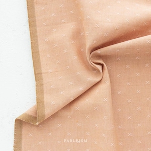 May include: A close-up of a peach-colored fabric with a white cross-stitch pattern. The fabric is folded and has a gold-colored edge.