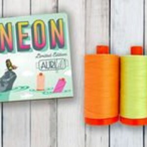 May include: Three spools of neon thread in orange, yellow, and pink, with orange caps, sit next to a box with the word "NEON" in large letters. The box has a light green background with illustrations of thread spools.