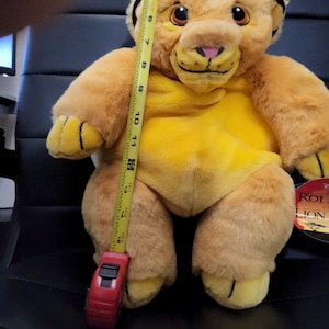 May include: A plush Simba toy from The Lion King, measuring approximately 15 inches tall. The toy is primarily yellow and tan with a pink nose and a tuft of hair. A measuring tape is held up against the toy.