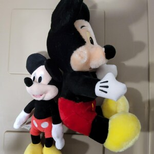May include: Two Mickey Mouse plush toys. The larger one is black, red, and yellow, with white gloves. The smaller one has the same color scheme. Both have black ears and smiling faces. The toys are soft and cuddly.