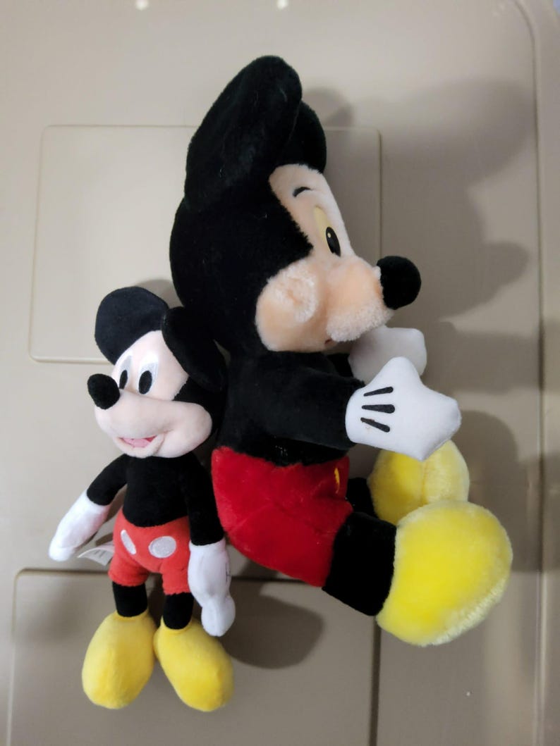 May include: Two Mickey Mouse plush toys. The larger one is black, red, and yellow, with white gloves. The smaller one has the same color scheme. Both have black ears and smiling faces. The toys are soft and cuddly.
