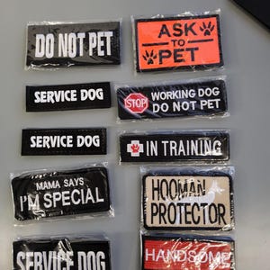 May include: A collection of black and colored patches with white text, including "DO NOT PET", "SERVICE DOG", "I'M SPECIAL", and "HANDSOME BOY". Some patches are in plastic packaging.