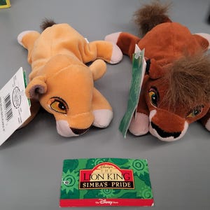 May include: Two plush lion cubs from Disney's The Lion King, one orange and one brown, with tags attached. The cubs are lying down, with their paws and bellies visible. A Simba's Pride card is at the bottom.
