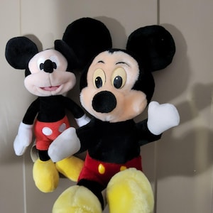 May include: Two plush Mickey Mouse toys. The toys have black fur, large black ears, white gloves, and yellow shoes. One Mickey Mouse is slightly smaller than the other. Both have red shorts with yellow buttons and a pink face.