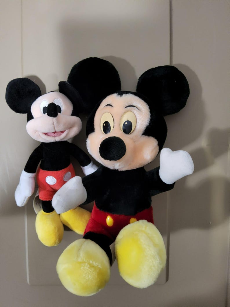 May include: Two plush Mickey Mouse toys. The toys have black fur, large black ears, white gloves, and yellow shoes. One Mickey Mouse is slightly smaller than the other. Both have red shorts with yellow buttons and a pink face.