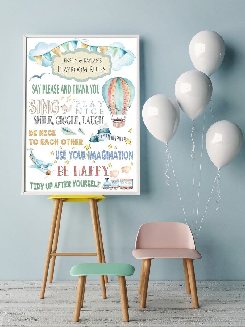 Op de afbeelding: Een ingelijste print met speelkamerregels, met aquarel illustraties van een luchtballon, vliegtuig en trein. De tekst bevat zinnen als "Zeg alsjeblieft en bedankt" en "Wees blij". Witte ballonnen en kleurrijke stoelen staan op de achtergrond.