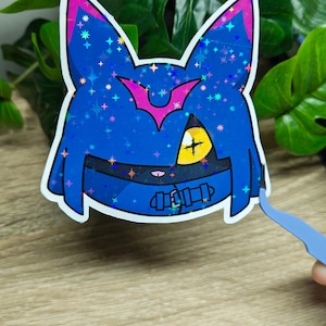 May include: A blue sticker featuring a cartoon character design. The character has a blue face with a pink and white outline, a yellow eye with a cross, and a black belt. The sticker has a holographic, glittery effect.