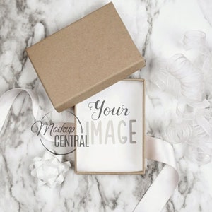 Könnte beinhalten: Eine braune Pappbox mit einer weißen Karte im Inneren. Die Karte trägt den Text "Your Image". Die Box steht auf einer weißen Marmorfläche mit weißem Band und einer weißen Schleife.