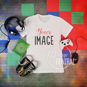 May include: A white t-shirt with the text "Your IMAGE" printed on the front. The t-shirt is lying flat on a colourful background with a pair of headphones, a game controller, a game disc, and a pair of trainers.