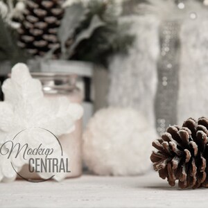 May include: A winter-themed still life featuring a brown pine cone, white snowflake ornament, and frosted greenery. The background includes a jar and other decorative elements, all set against a soft, neutral backdrop. The image has the text "Mockup CENTRAL".
