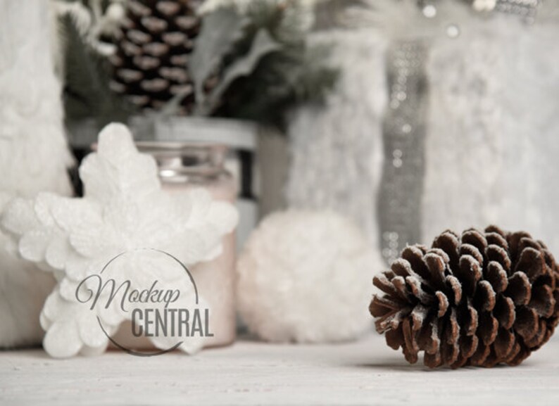 May include: A winter-themed still life featuring a brown pine cone, white snowflake ornament, and frosted greenery. The background includes a jar and other decorative elements, all set against a soft, neutral backdrop. The image has the text "Mockup CENTRAL".