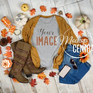 May include: A gray t-shirt with the text "Your Image" on it, a mustard yellow cardigan, blue jeans, brown boots, and fall-themed decorations on a white wood background.