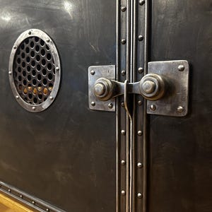 May include: Close-up of a black metal cabinet with two doors. The doors have a decorative metal trim with rivets and a latch with a round knob. The cabinet has a round vent with a metal grate.