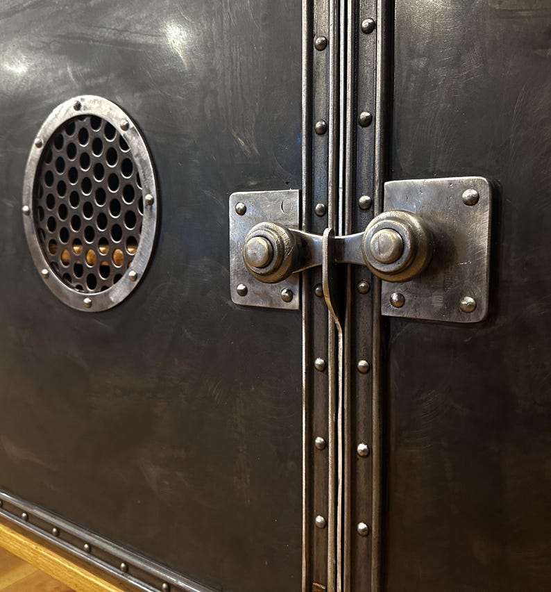 May include: Close-up of a black metal cabinet with two doors. The doors have a decorative metal trim with rivets and a latch with a round knob. The cabinet has a round vent with a metal grate.