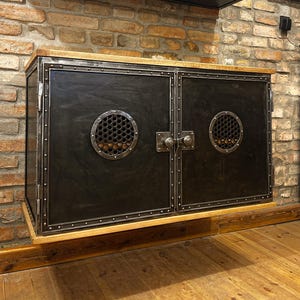 May include: A black metal cabinet with two doors and a wood top. The cabinet has two round metal vents with a honeycomb pattern. The cabinet is mounted on a wall.