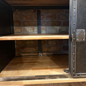 May include: A black metal cabinet with a wooden top and shelves. The cabinet has a single door with a metal handle and rivets. The interior of the cabinet is visible, showing a brick wall and a wooden shelf.