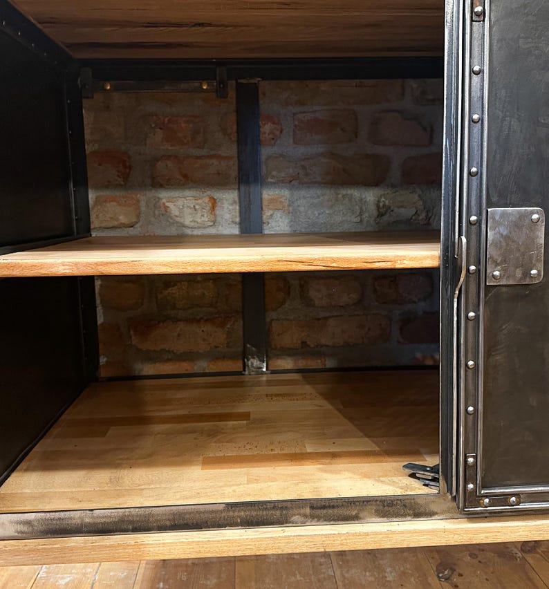 May include: A black metal cabinet with a wooden top and shelves. The cabinet has a single door with a metal handle and rivets. The interior of the cabinet is visible, showing a brick wall and a wooden shelf.