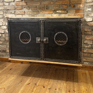 May include: A black metal cabinet with two doors and a wood top. Each door has a circular cutout with a metal grate. The cabinet is mounted on a brick wall.