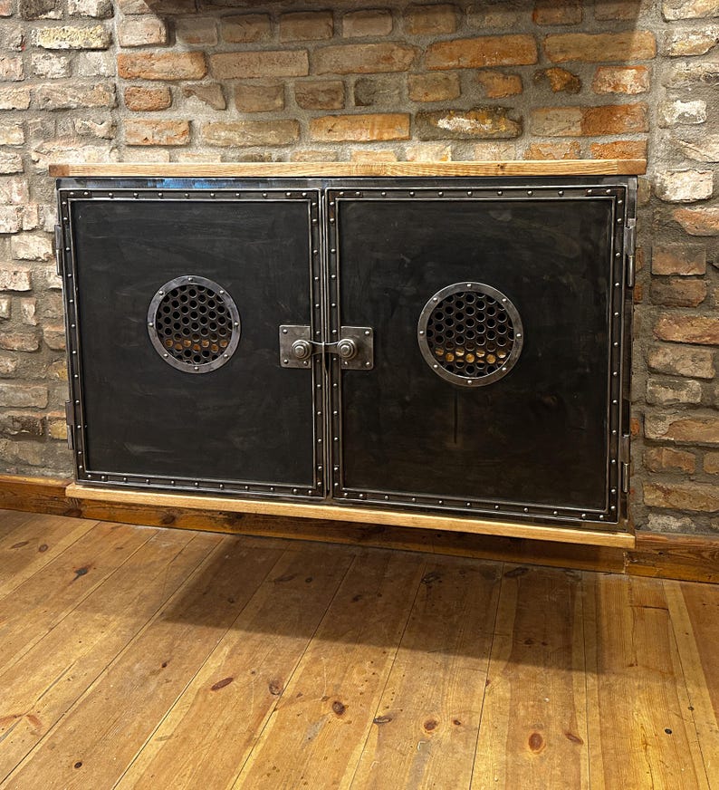 May include: A black metal cabinet with two doors and a wood top. Each door has a circular cutout with a metal grate. The cabinet is mounted on a brick wall.
