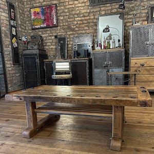 Könnte beinhalten: Eine große, rustikale Werkbank aus Holz mit einem unteren Regal und sichtbaren Verbindungen. Die Werkbank besteht aus verwittertem Holz und hat eine rechteckige Oberseite mit mehreren Löchern. Die Beine sind dick und stabil, und das Gesamtdesign deutet auf eine Vintage- oder Industrieästhetik hin.