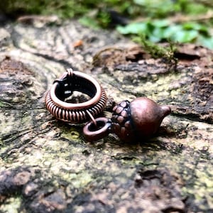 May include: A copper acorn charm and a small, round, openwork charm with a ring, on a textured, brown surface. The acorn charm has a detailed cap and a smooth body. The background is blurred with green foliage and moss.