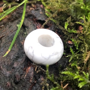May include: A white, round bead with a hole in the center. The bead is made of a natural material, possibly stone or bone. It is sitting on a bed of moss and dirt.